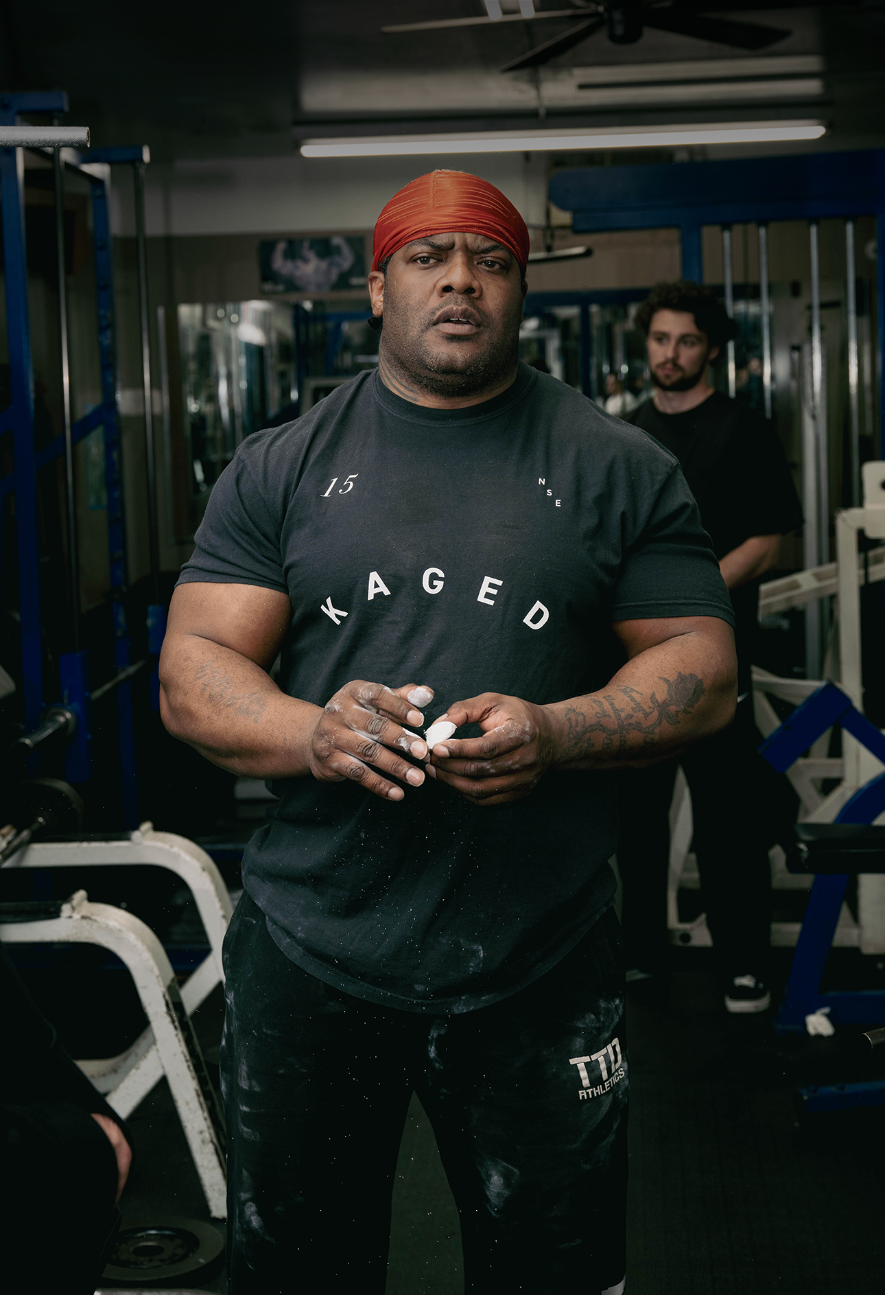 Muscular man standing in a gym wearing a black KAGED T-shirt and red headwrap, holding chalk with weight equipment behind him