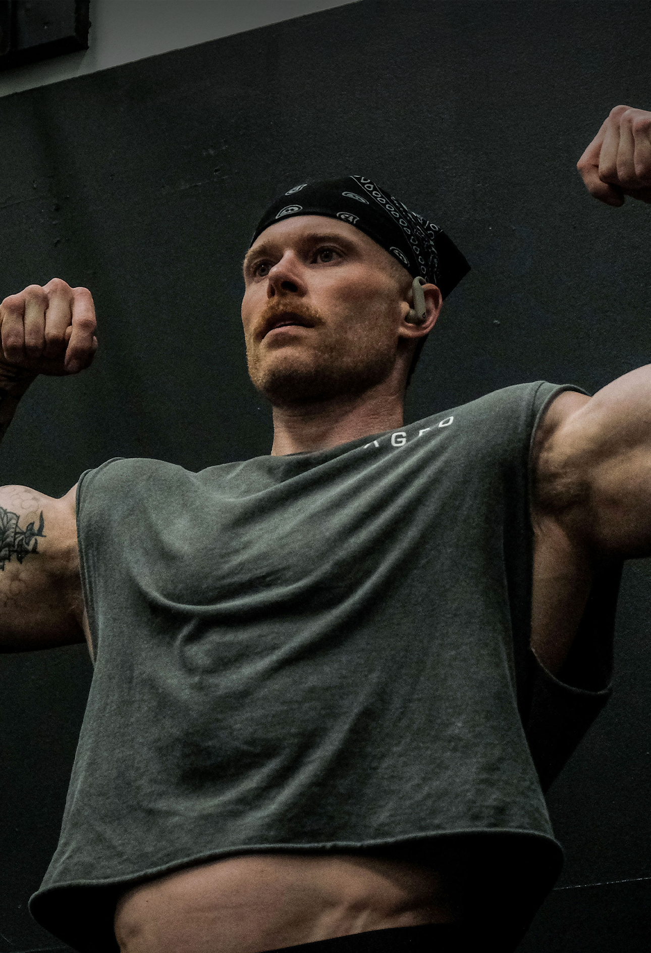 Man flexing in a gym while wearing a dark sleeveless KAGED shirt and black bandana